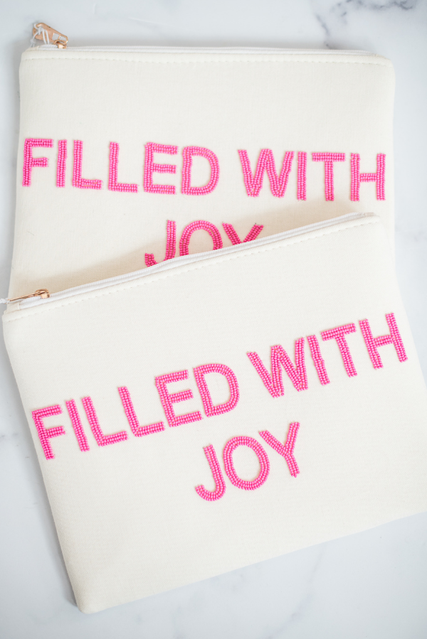 two white canvas pouches with hot pink block letters saying "filled with joy" with gold zipper and white marble background