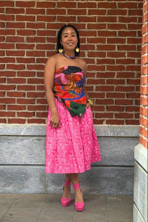 Woman in a colorful strapless top and pink skirt standing against a brick wall.