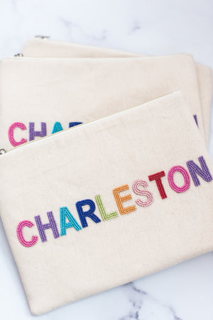 white canvas pouch with rainbow embellishments spelling out "charleston" with hot pink, purple, teal, blue, lime green, orange, light pink, and red beaded letters on white marble background