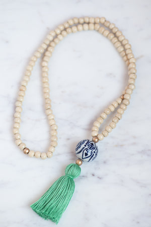 Model wearing wood beaded necklace with gold accents, porcelain chinoiserie statement bead, and kelly green tassel. 